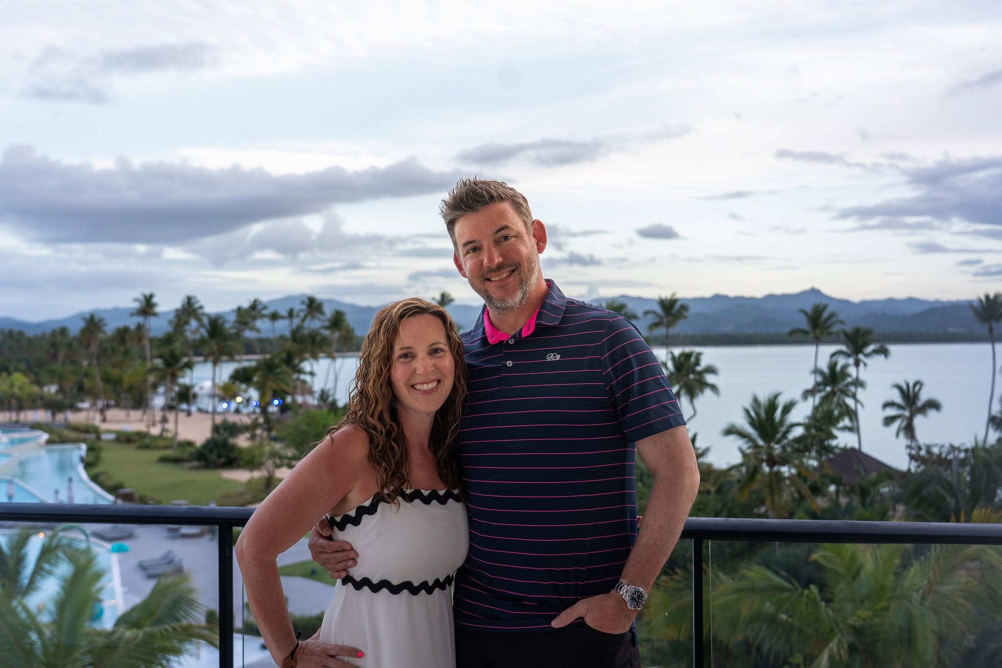 Robb Sutton and his wife on a balcony overlooking a beach, pool and palm trees
